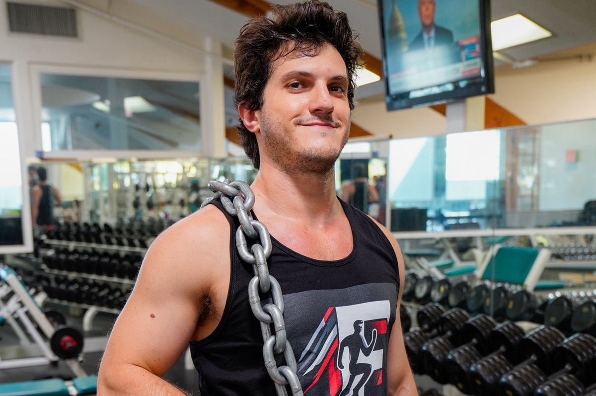 Jamin Fitness founder demonstrating strength with heavy chains in gym, wearing branded black tank top and red shorts