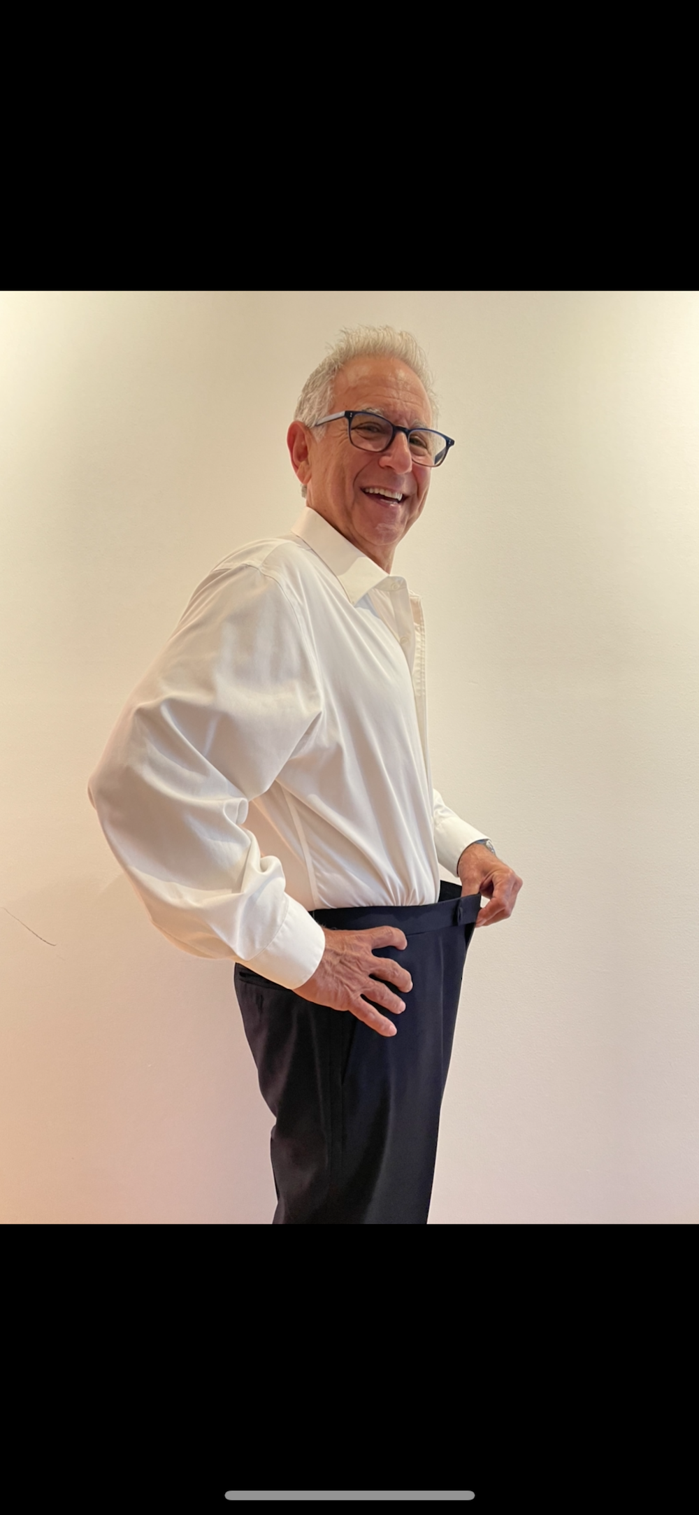 Senior fitness transformation success story showing client in white dress shirt demonstrating significant weight loss results by pulling loose-fitting navy pants, highlighting personal training effectiveness