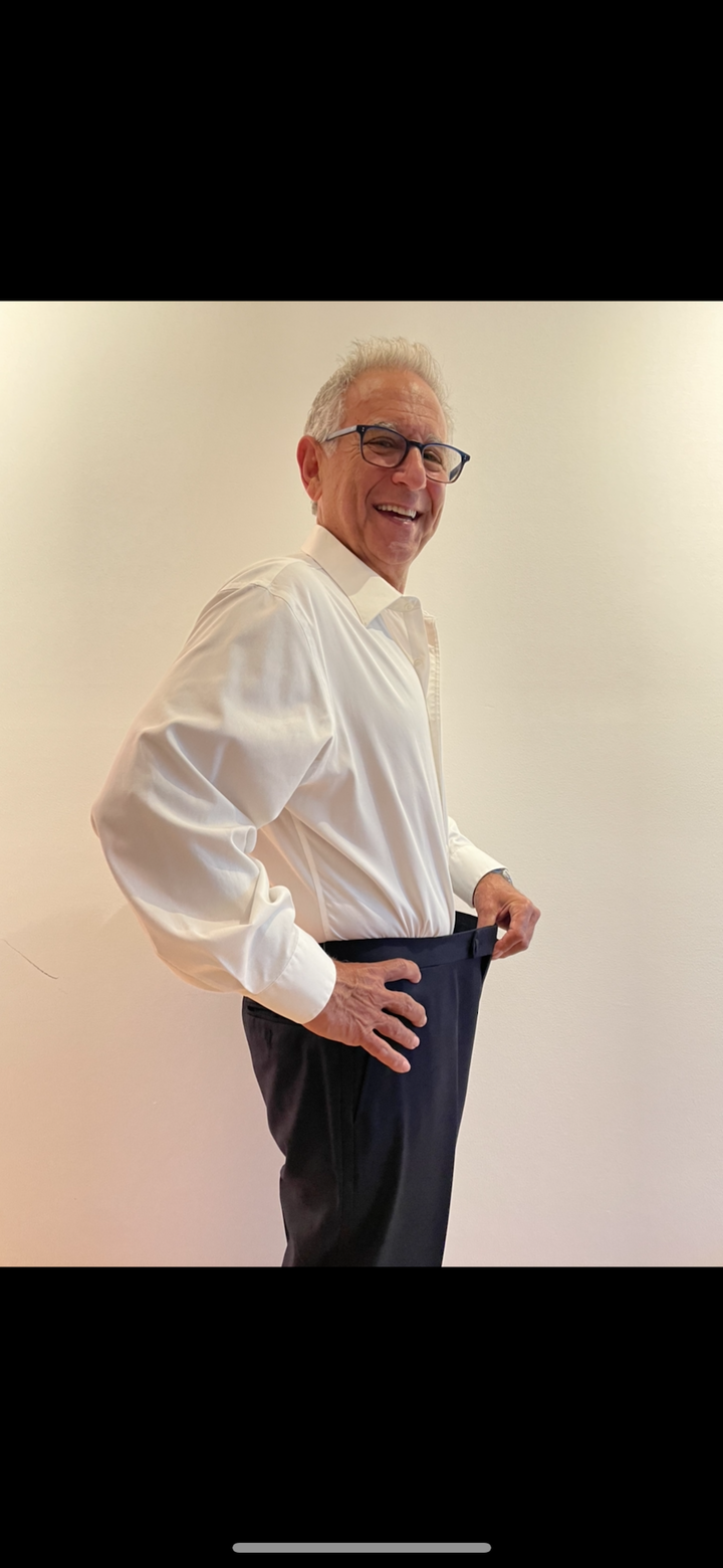 Senior fitness transformation success story showing client in white dress shirt demonstrating significant weight loss results by pulling loose-fitting navy pants, highlighting personal training effectiveness