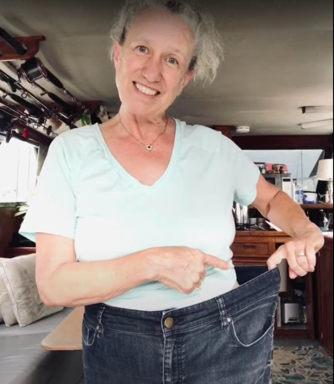 Weight loss success story showing woman in light blue v-neck shirt demonstrating significant progress by holding out loose-fitting jeans, showcasing transformation results in home setting with genuine smile